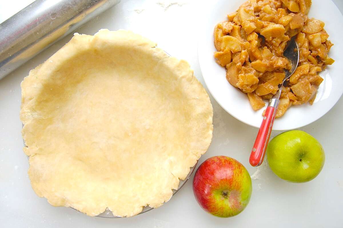 Rolled out pie crust in a pan alongside fresh apples and a bowl of apple pie filling.